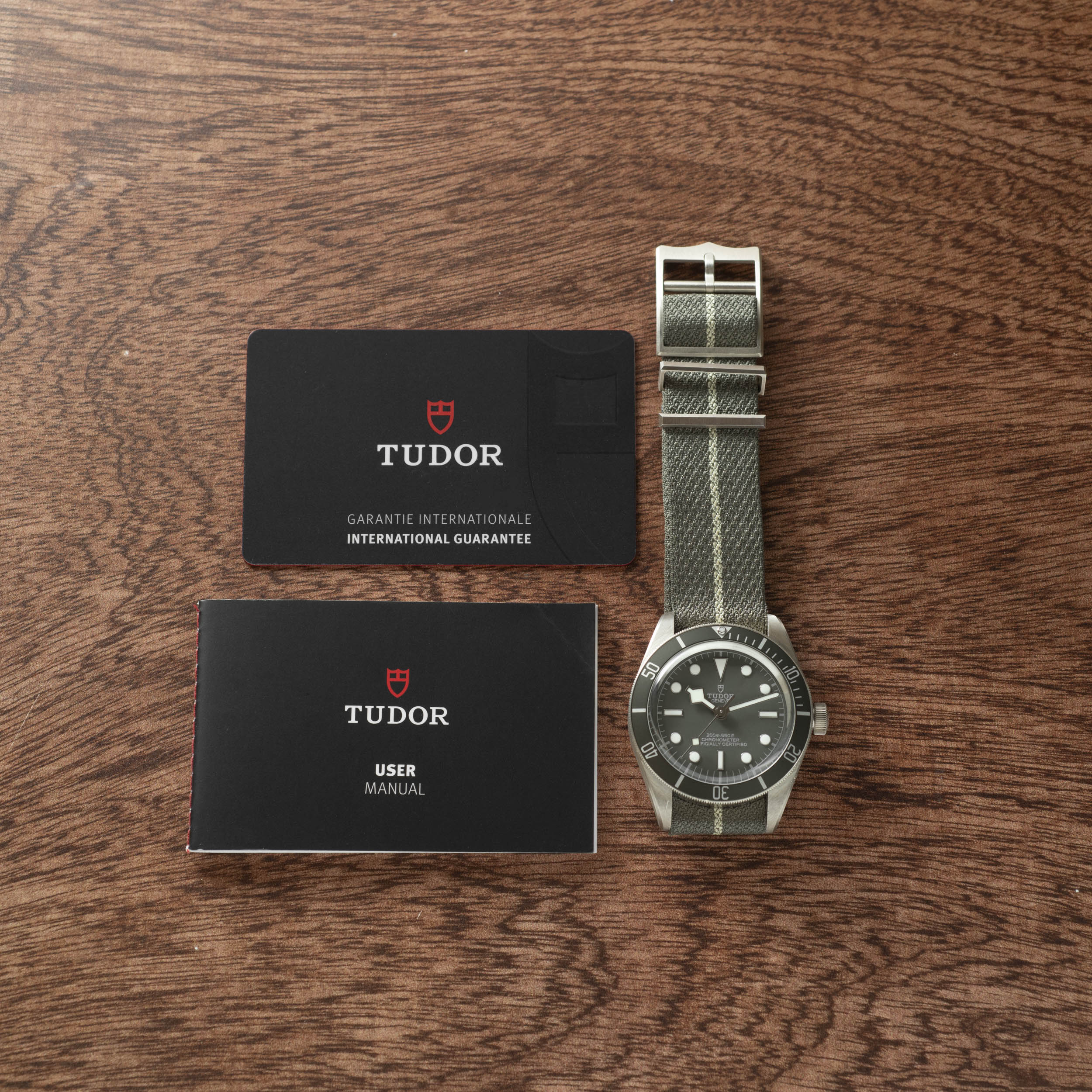 A TUDOR Black Bay 58 Silver watch with a striped strap, warranty card, and user manual displayed on a wooden surface.