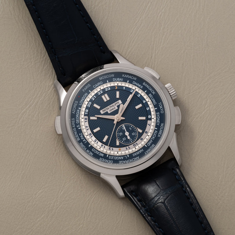 The Patek Philippe World Time Chronograph, featuring a blue dial with world city names and a dark leather strap, rests on beige leather.