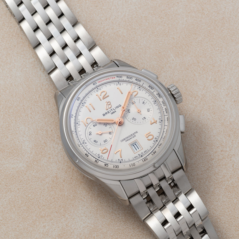 A Breitling B01 Premier Chronograph 42 wristwatch with a silver metal band, white dial, and rose gold-tone hour markers displayed on a beige surface.