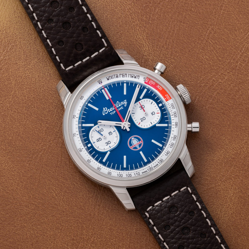 A Breitling Top Time B01 Chronograph Shelby Cobra wristwatch with a blue dial and black leather strap is displayed on a brown leather surface.