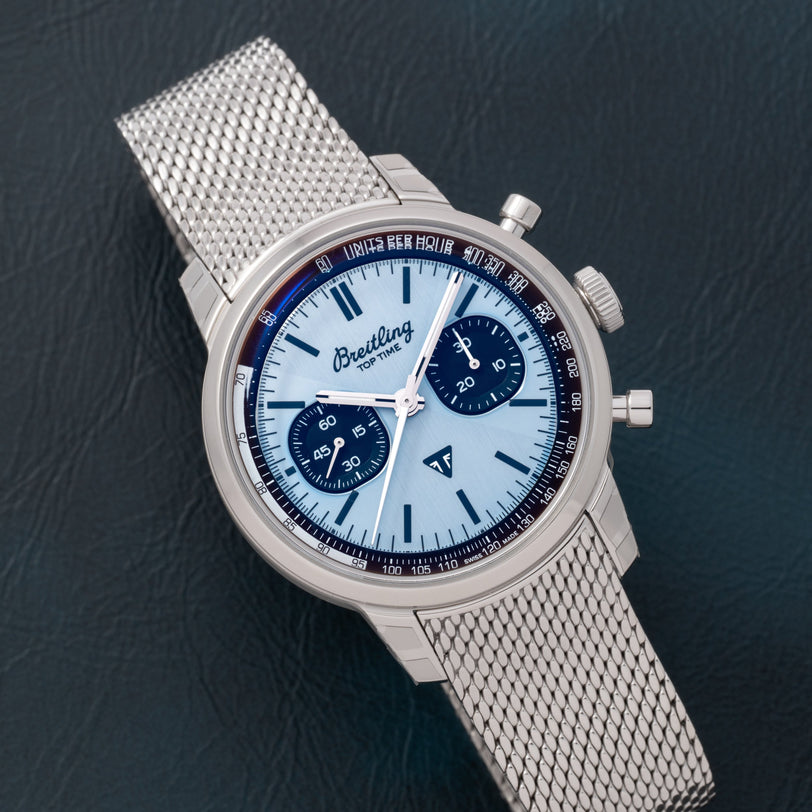 A Breitling Top Time B01 Chronograph Triumph watch with a silver mesh band and light blue dial is displayed on a dark textured surface.