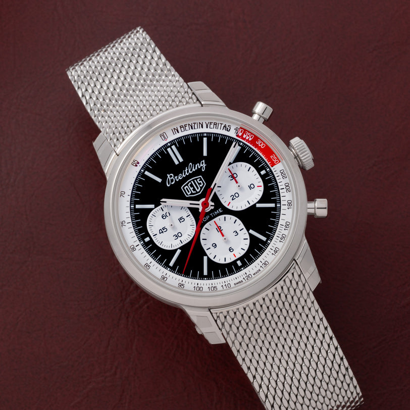 A Breitling Top Time B01 Chronograph Deus wristwatch with a mesh strap rests on a brown leather surface.