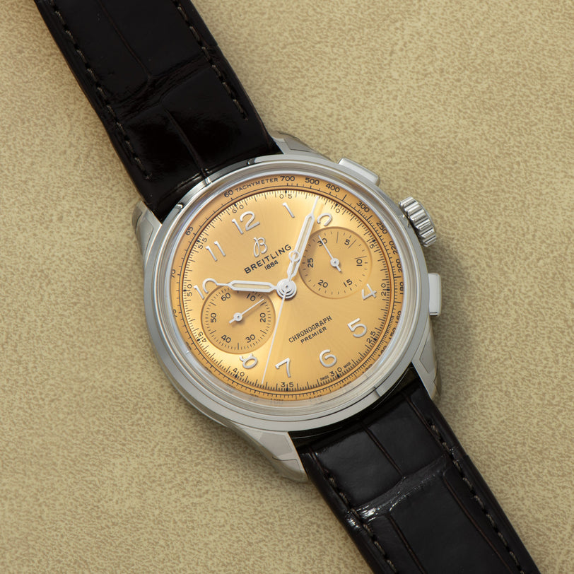 The Breitling Premiere B09 Chronograph 40 features a gold dial, black leather strap, and silver case, displayed on a beige surface.