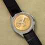 The Breitling Premiere B09 Chronograph 40 features a gold dial, black leather strap, and silver case, displayed on a beige surface.