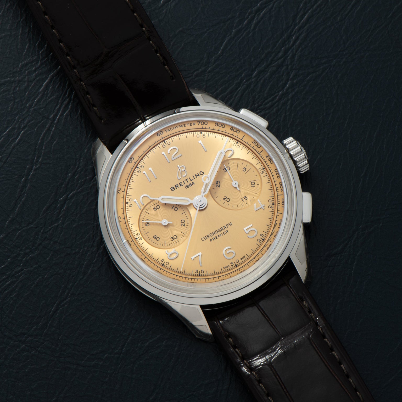 The Breitling Premiere B09 Chronograph 40, featuring a tan dial and black leather strap, is displayed on a dark textured surface.