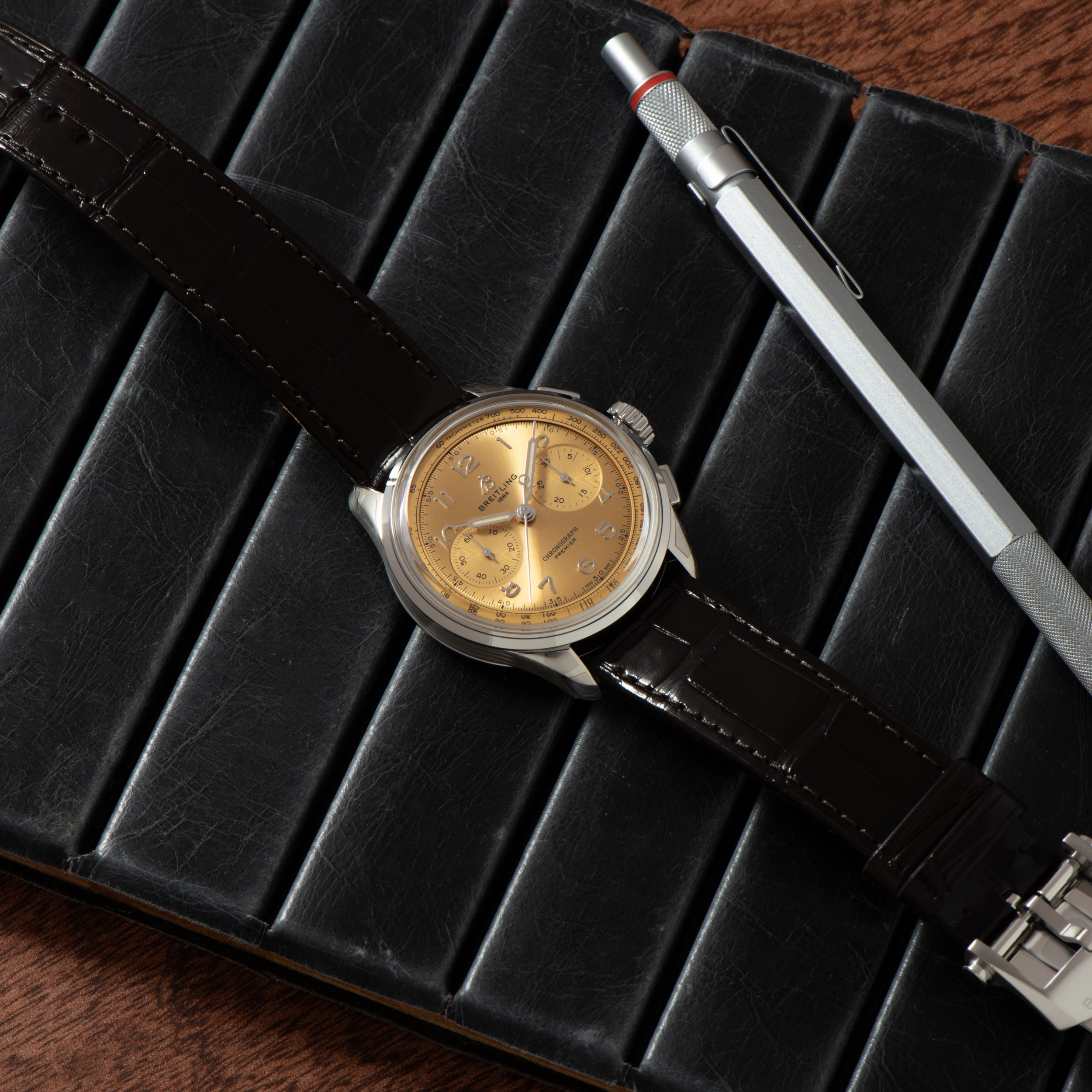 The Breitling Premiere B09 Chronograph 40, featuring a gold face and dark leather strap, is displayed on a black notebook next to a silver pen.
