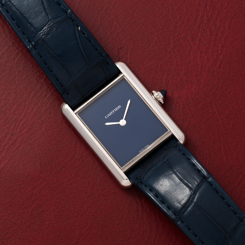 A Cartier Tank Must watch with a blue square face and leather strap rests on a textured red surface.