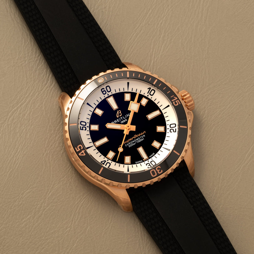The Breitling Superocean Automatic 42, a gold-tone dive watch with a black dial and black strap, rests on a beige surface.