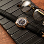 Breitling Superocean Automatic 42 wristwatch with black strap and eyeglasses resting on a black leather pad atop a wooden surface.