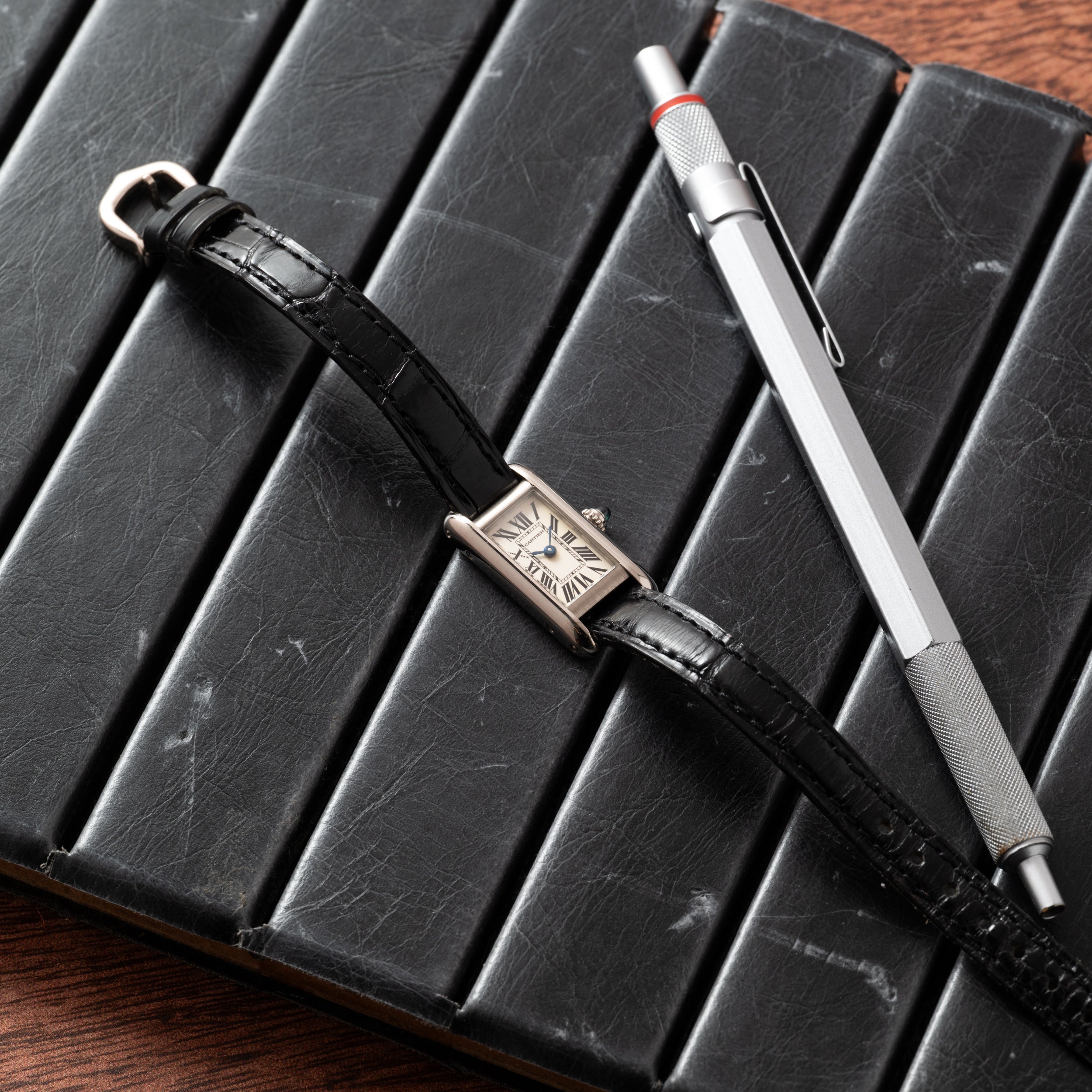 A Cartier Tank Ladies Allongee watch with a black leather strap and a silver pen placed on a black notebook.