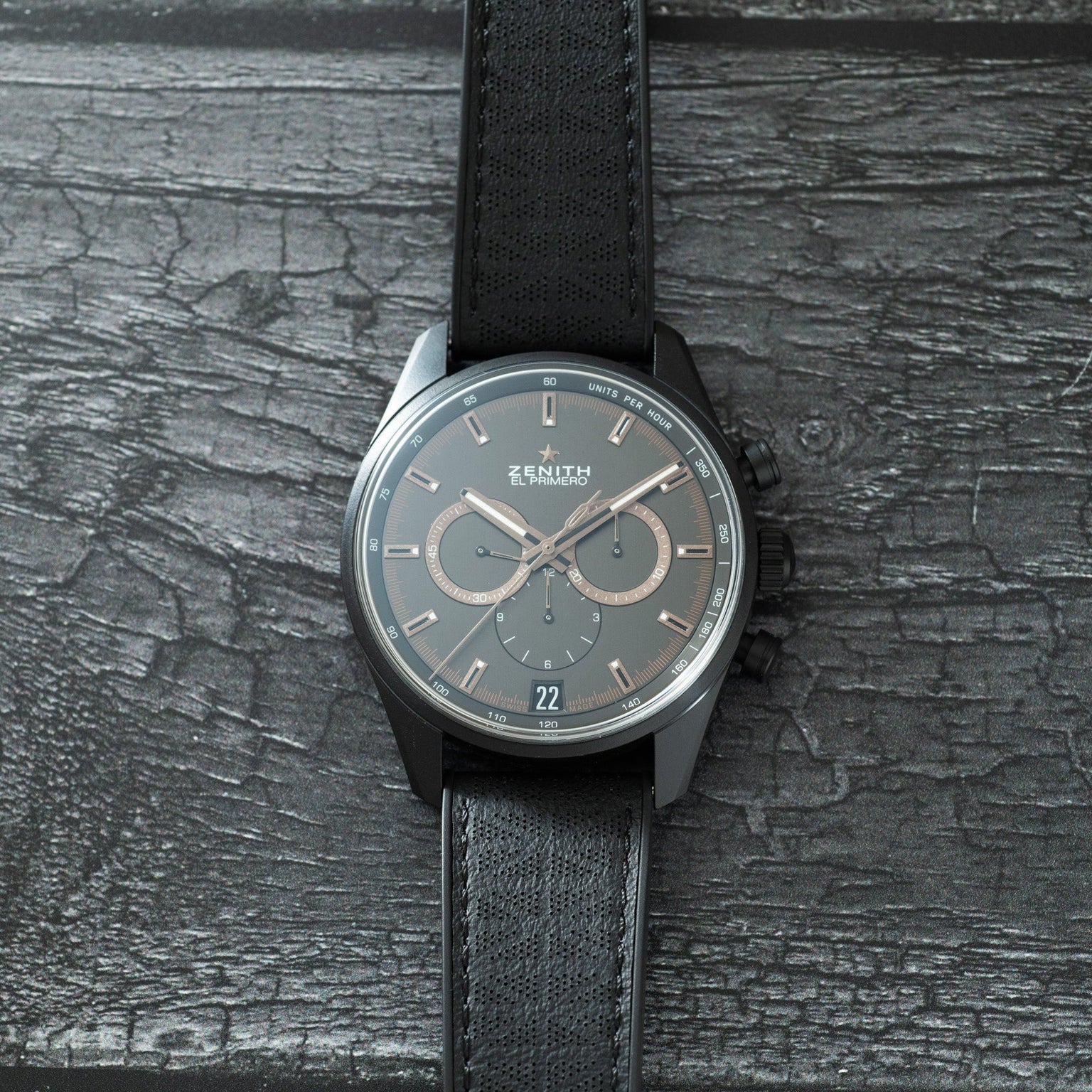 Zenith chronograph watch with dark gray dial, three subdials, date window, and black textile strap on textured surface.