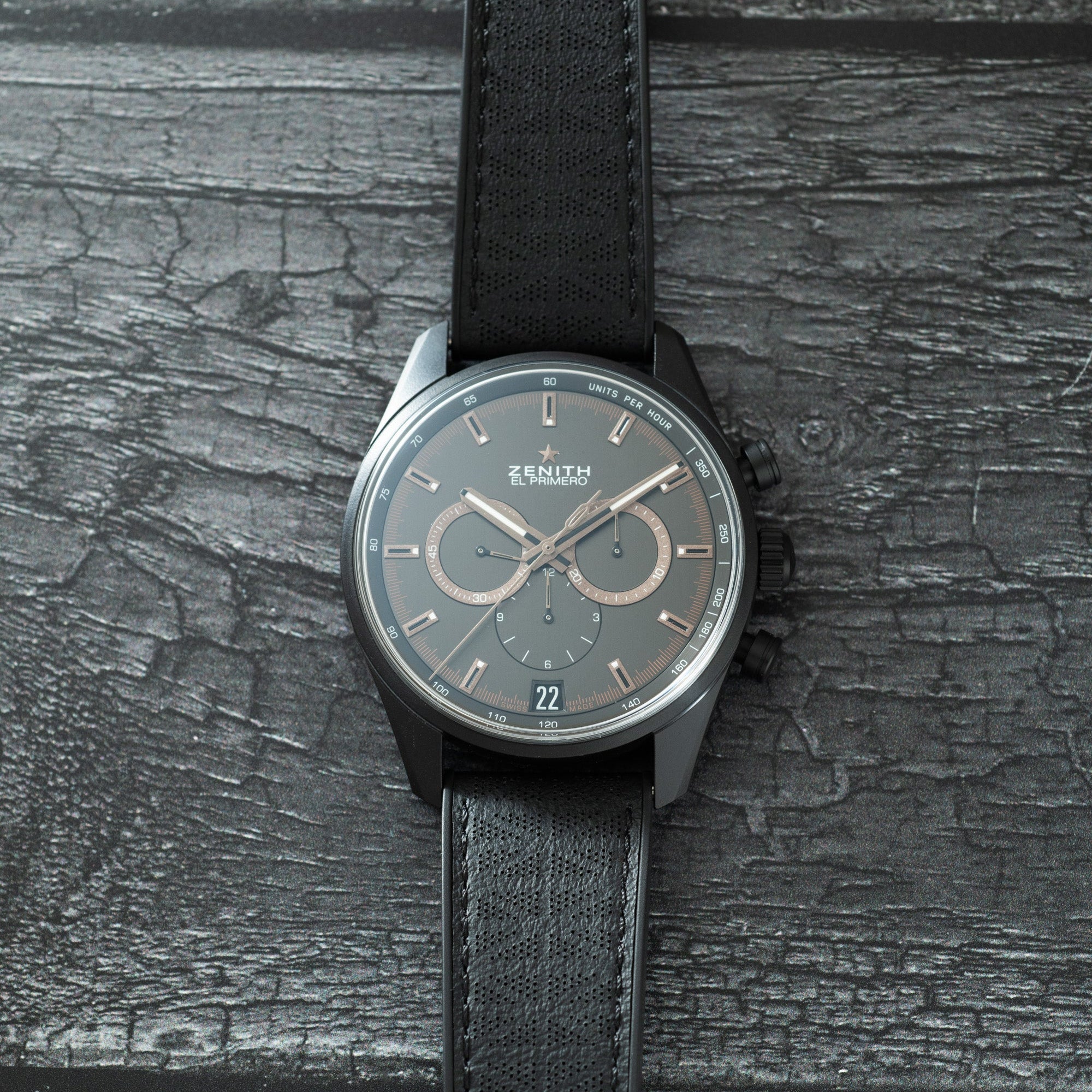 Zenith chronograph watch with dark gray dial, three subdials, date window, and black textile strap on textured surface.