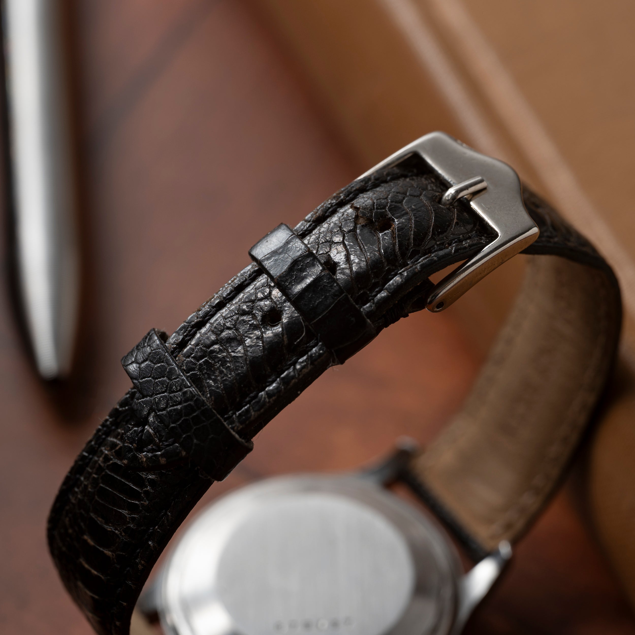 Close-up of a Jaeger LeCoultre 'European Market' Powermatic midcentury watch with a textured black leather strap and silver buckle on wood. Focus is on the strap; watch face and a metal pen appear blurred in the background.
