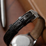 Close-up of a Jaeger LeCoultre 'European Market' Powermatic midcentury watch with a textured black leather strap and silver buckle on wood. Focus is on the strap; watch face and a metal pen appear blurred in the background.