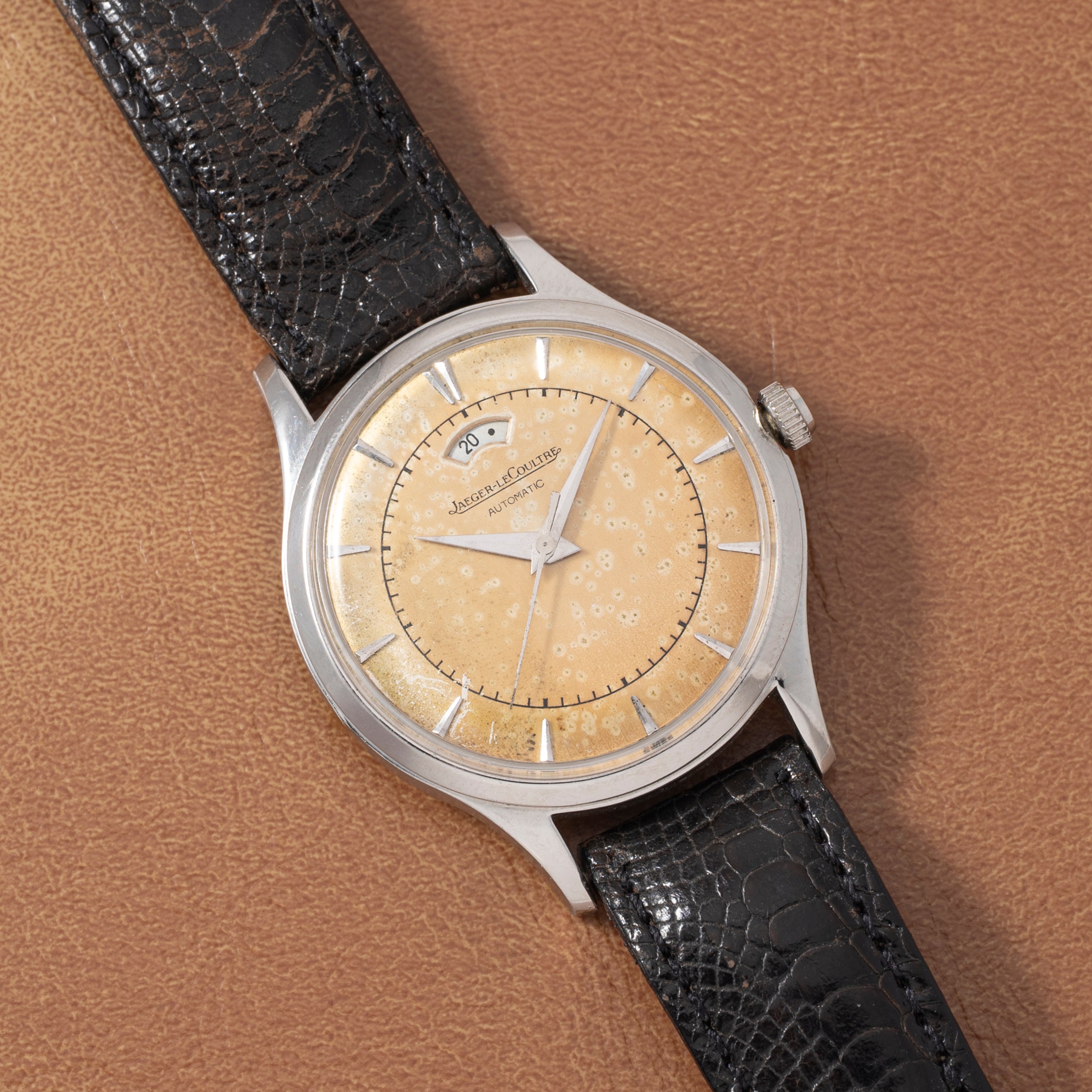 A Jaeger LeCoultre 'European Market' Powermatic wristwatch with a patinated beige dial, silver indices, date window, power-reserve display, and black leather strap is showcased on a brown textured surface.