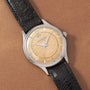 A Jaeger LeCoultre 'European Market' Powermatic wristwatch with a patinated beige dial, silver indices, date window, power-reserve display, and black leather strap is showcased on a brown textured surface.