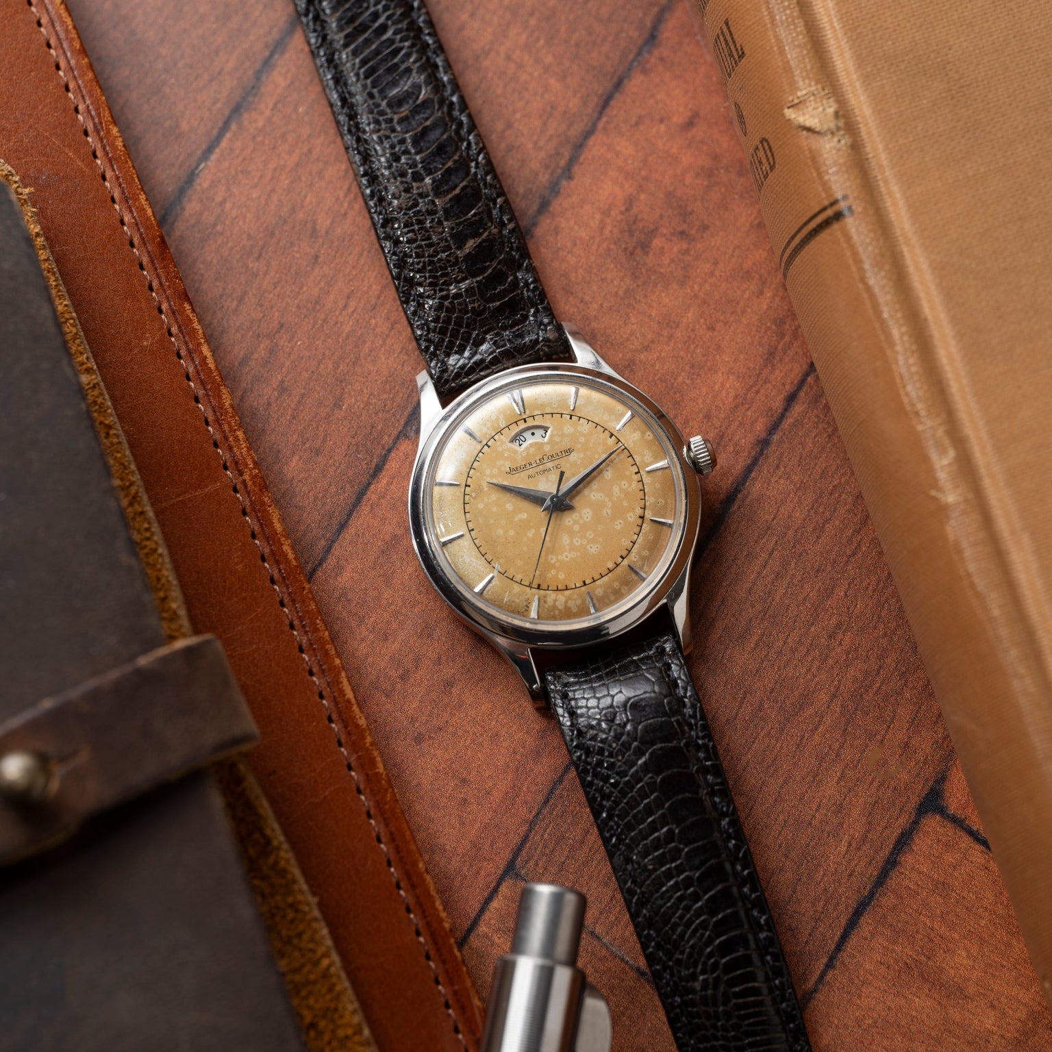 A Jaeger LeCoultre 'European Market' Powermatic wristwatch with a beige, weathered dial and black leather strap sits on a wooden surface among leather-bound books and a pen—an iconic midcentury dress watch.