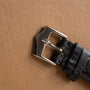 Close-up of the Jaeger-LeCoultre 'European Market' Powermatic's silver metal buckle on a black leather strap, resting on a textured light brown surface.