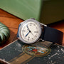 The Movado 'Calatrava' Steel, a vintage Swiss watch with a cream dial, Arabic numerals, striking blue second hand, and black leather strap, rests on a worn book near a green decorative object on a wooden surface.