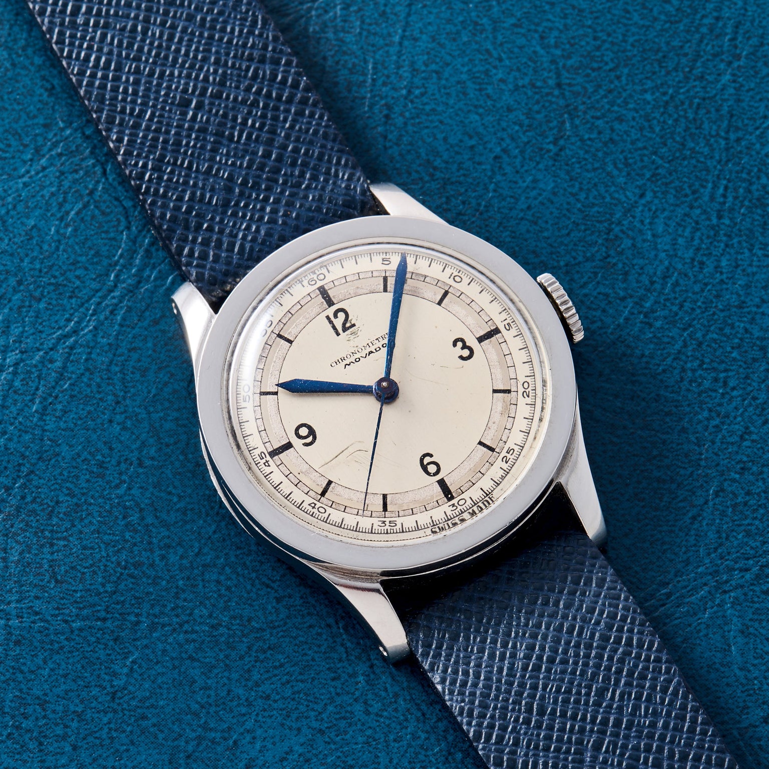The Movado ‘Calatrava’ Steel watch, with a cream dial, blue hands, and black numerals on a blue textured leather strap, is displayed atop a matching blue textured surface.