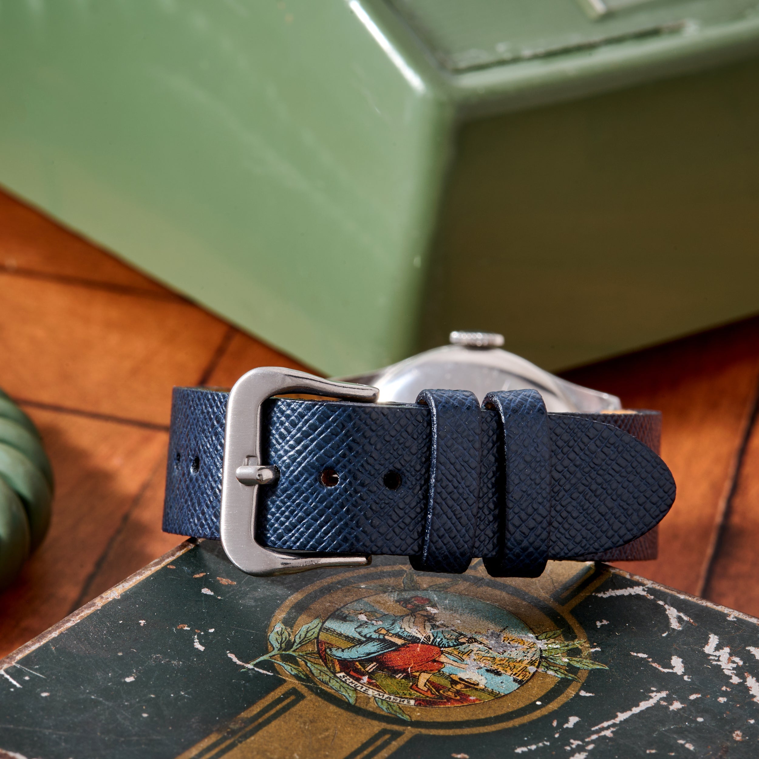 A close-up of a blue textured leather strap with a silver buckle, attached to the Movado 'Calatrava' Steel watch, rests on a vintage book; in the background, a green metal box evokes Swiss watchmaking elegance.