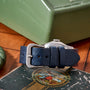 A close-up of a blue textured leather strap with a silver buckle, attached to the Movado 'Calatrava' Steel watch, rests on a vintage book; in the background, a green metal box evokes Swiss watchmaking elegance.