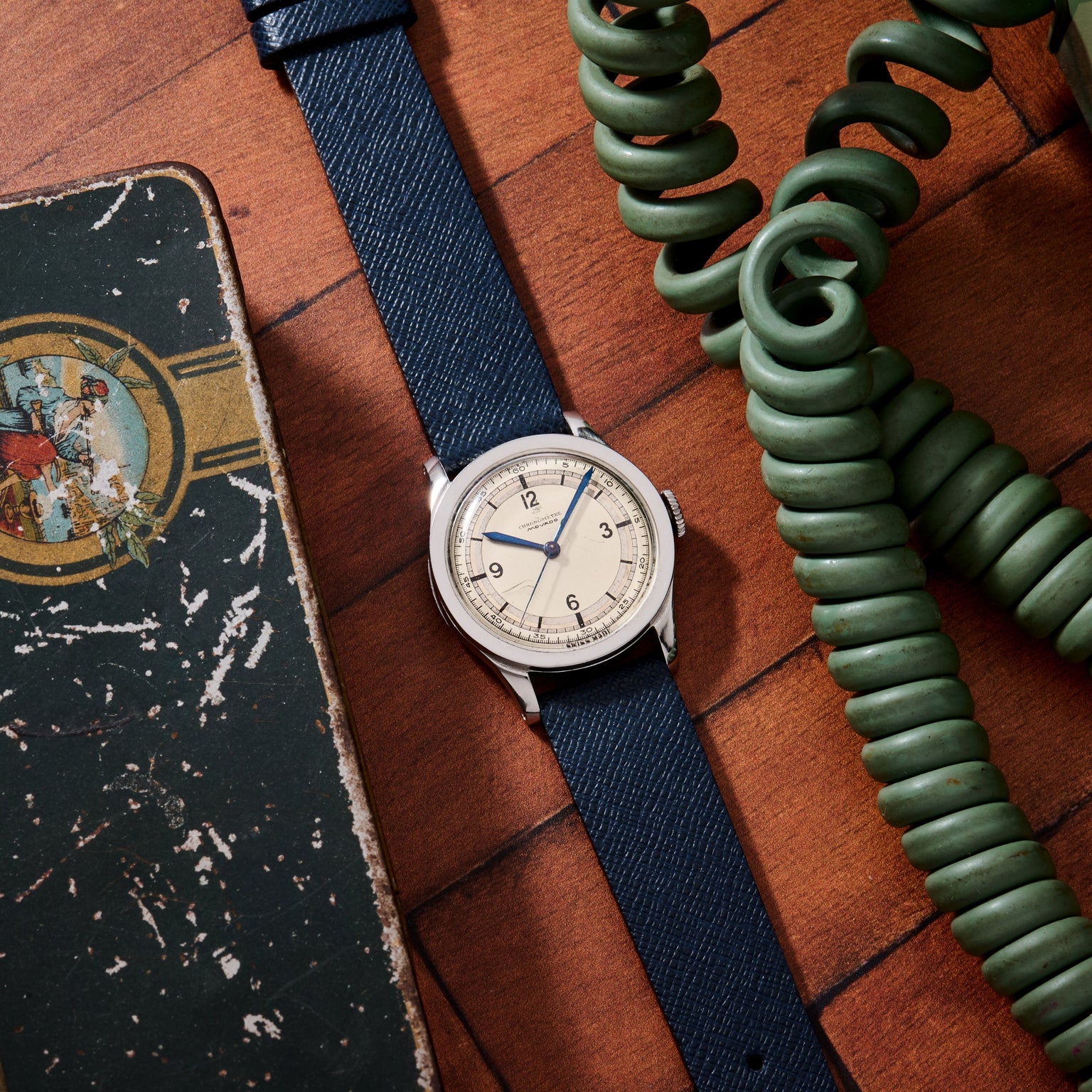The Movado 'Calatrava' Steel watch with a navy strap rests on a wooden surface beside a green coiled phone cord and a worn illustrated box, capturing the timeless charm of Swiss watchmaking.
