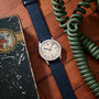 The Movado 'Calatrava' Steel watch with a navy strap rests on a wooden surface beside a green coiled phone cord and a worn illustrated box, capturing the timeless charm of Swiss watchmaking.