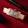 A rectangular ladies' Cartier Tank Americaine in 18K rose gold with a silver guilloché dial and Roman numerals lies on red fabric, with portions of a black pen and leather case visible nearby.