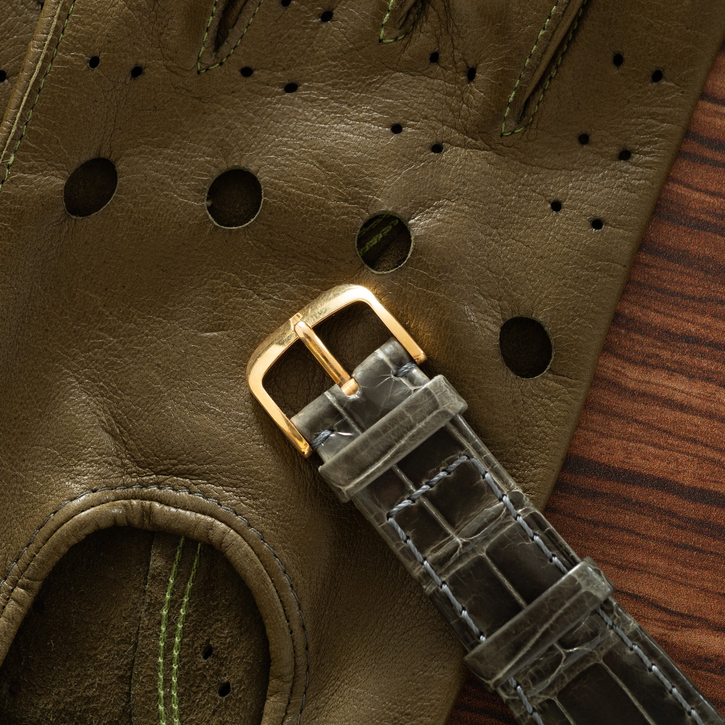 Gray crocodile leather watch strap with gold-tone buckle on olive perforated leather glove.