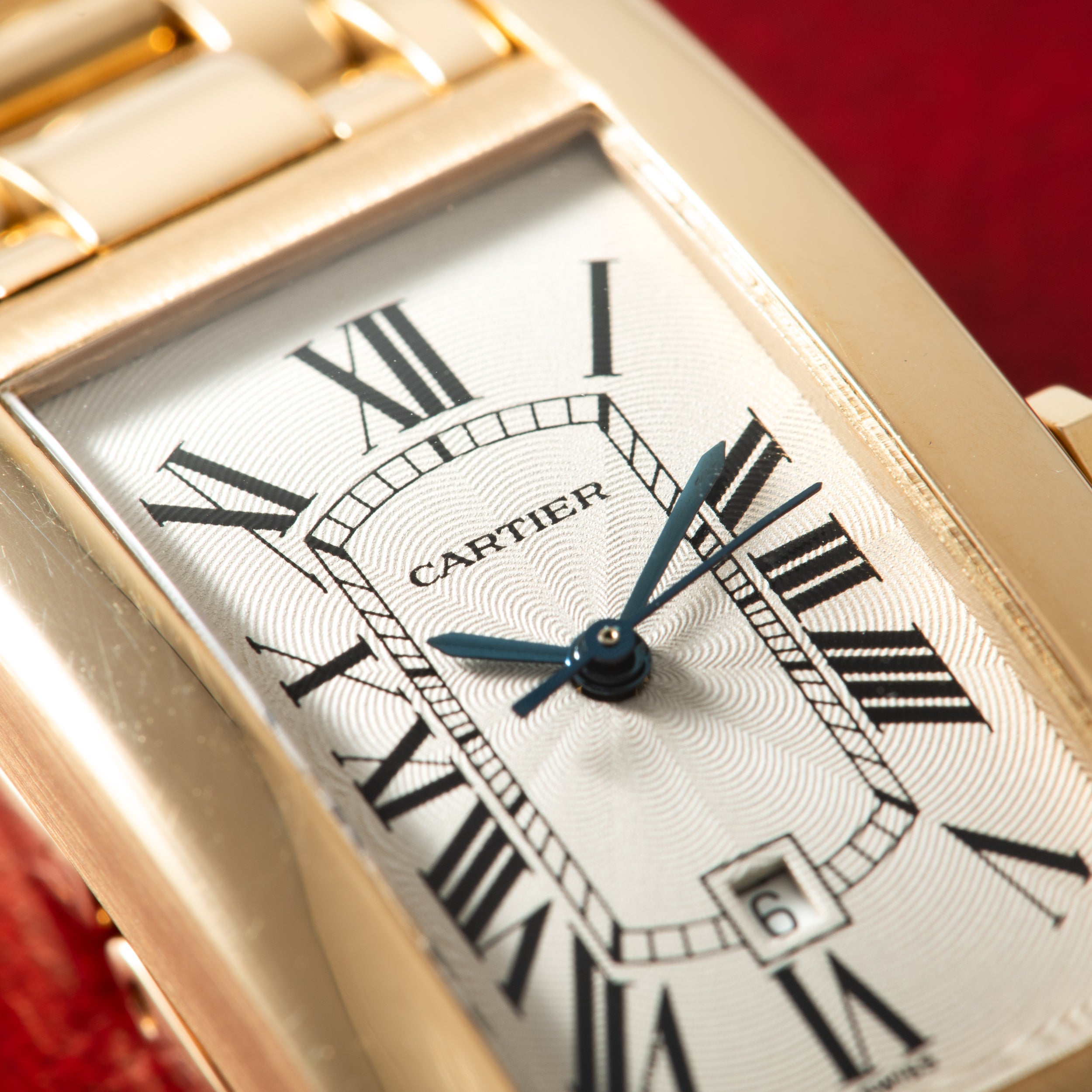 Gold Cartier Tank Américaine watch with white dial, Roman numerals, and blue hands on red background.