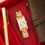 Gold-tone rectangular watch with white dial and Roman numerals on red fabric next to a pen and leather accessories.