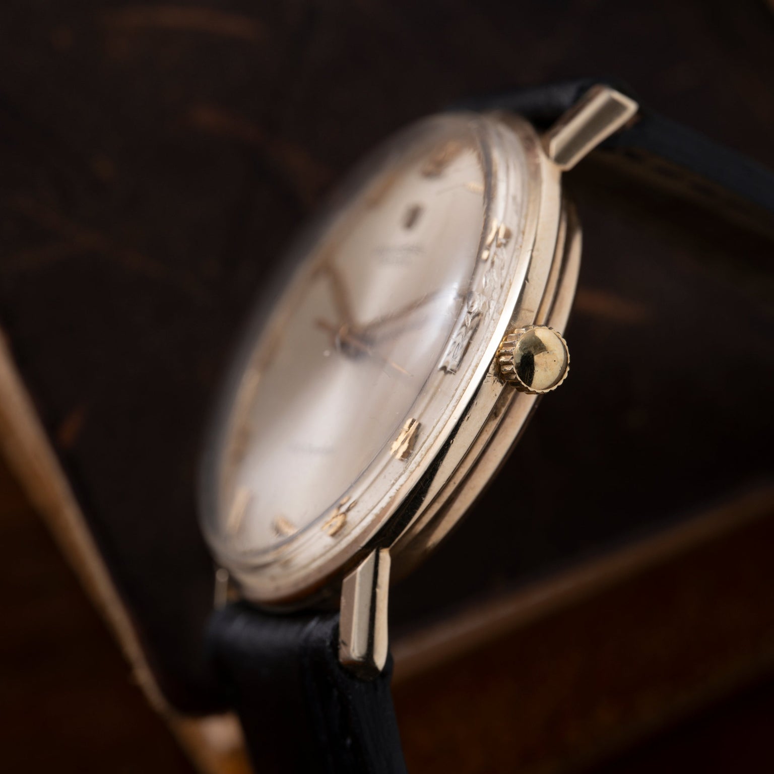 Gold-tone round watch with cream dial, Arabic numerals, and black leather strap displayed at an angle.
