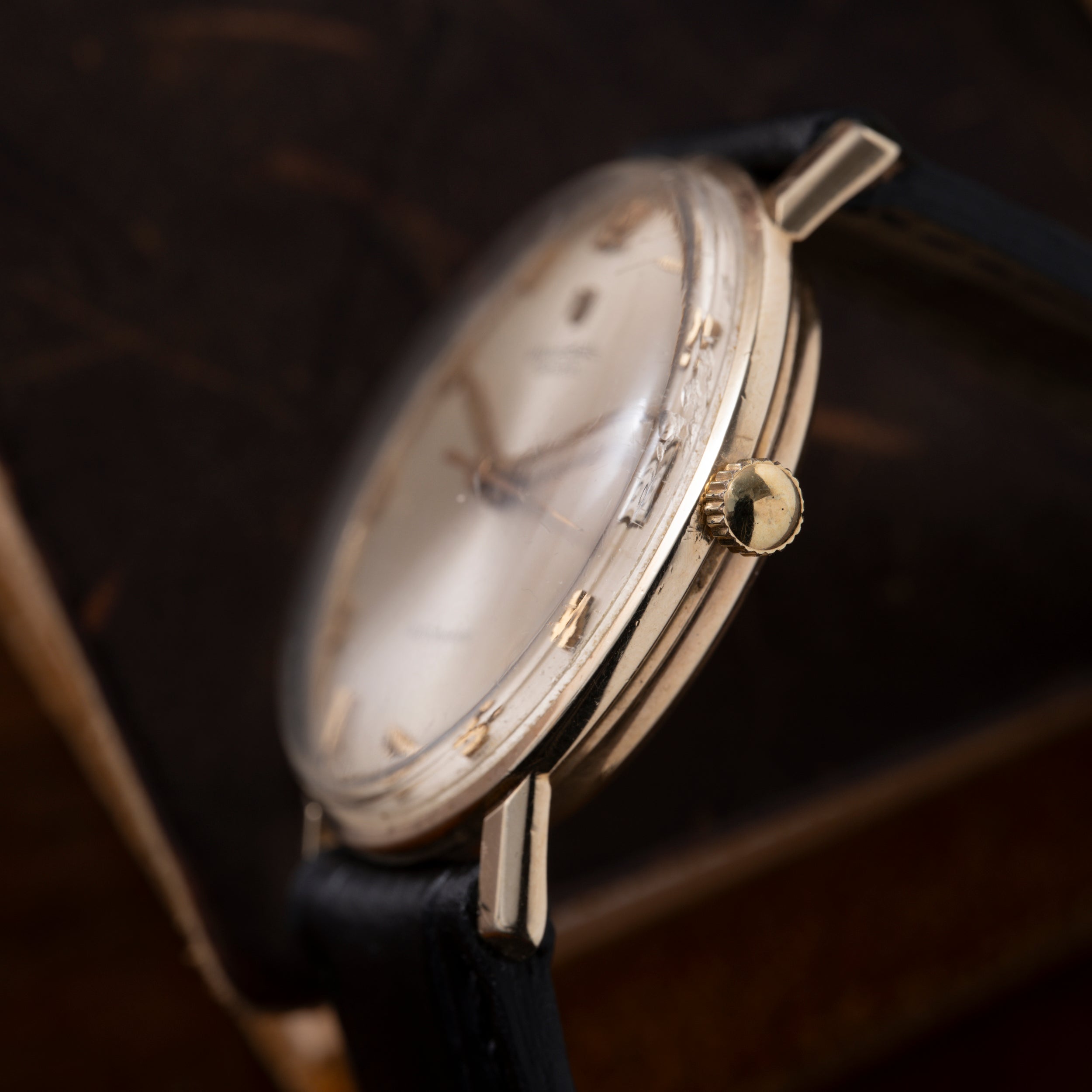 Gold-tone round watch with cream dial, Arabic numerals, and black leather strap displayed at an angle.