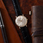 Gold-tone Universal Geneve watch with silver dial, black leather strap, and date window on brown leather background.