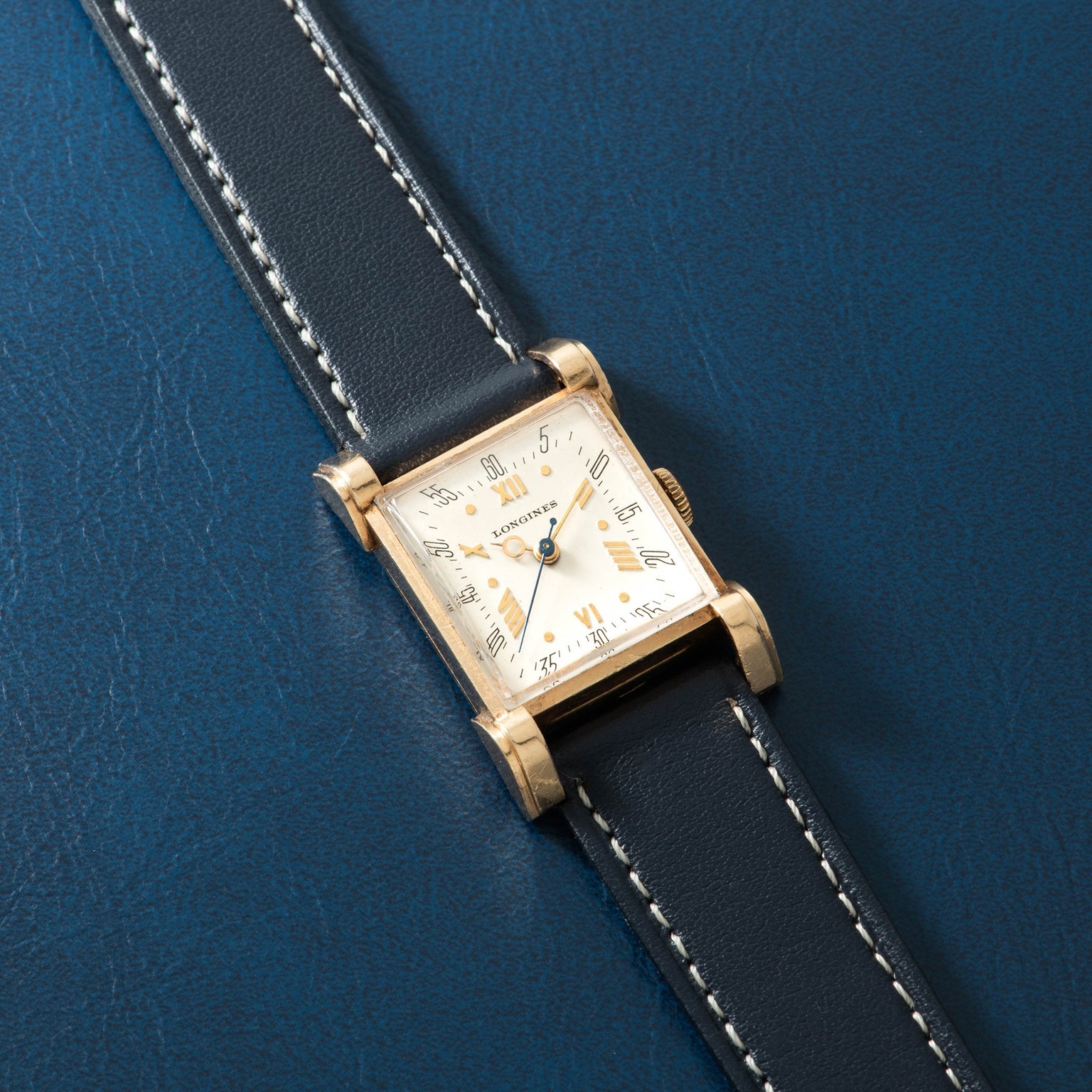 Gold-tone rectangular watch with cream dial, Arabic numerals, and black leather strap with white stitching on blue background