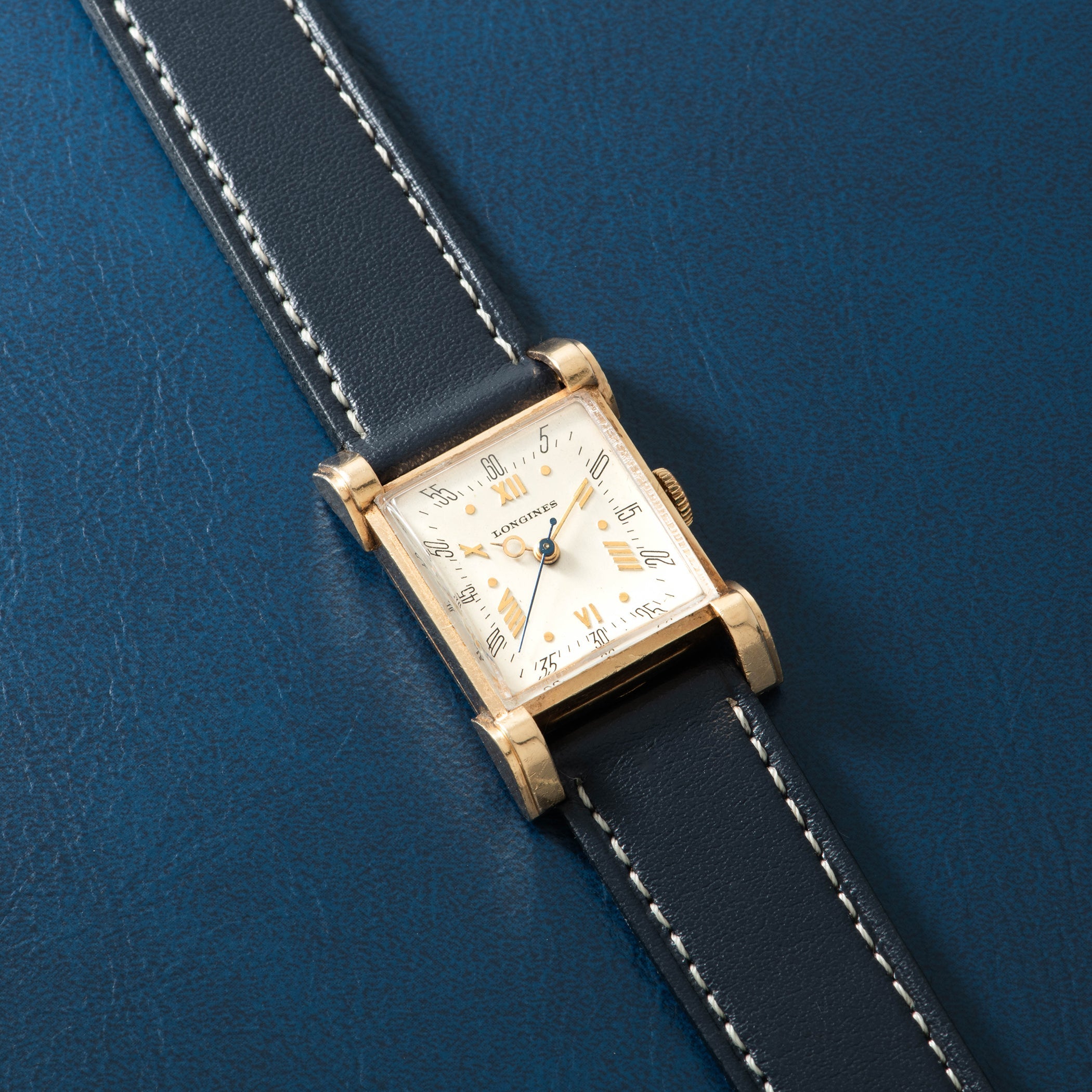 Gold-tone rectangular watch with cream dial, Arabic numerals, and black leather strap with white stitching on blue background