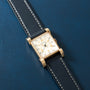 Gold-tone rectangular watch with cream dial, Arabic numerals, and black leather strap with white stitching on blue background
