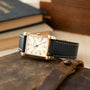 Longines Tank watch with rectangular gold case, cream dial with Roman numerals, and black leather strap with white stitching.