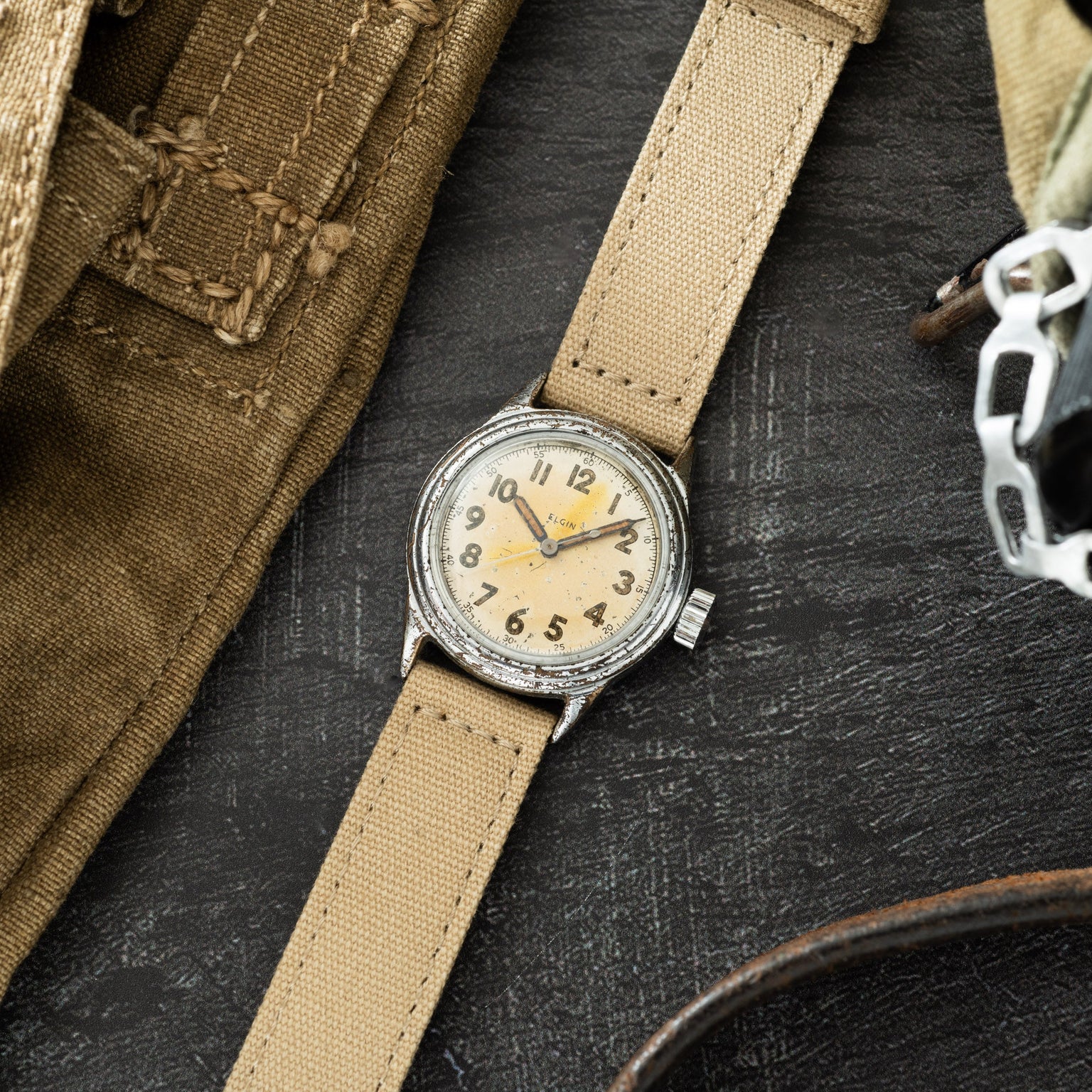 Elgin watch with cream dial and Arabic numerals on tan canvas strap, displayed on dark surface with military gear.