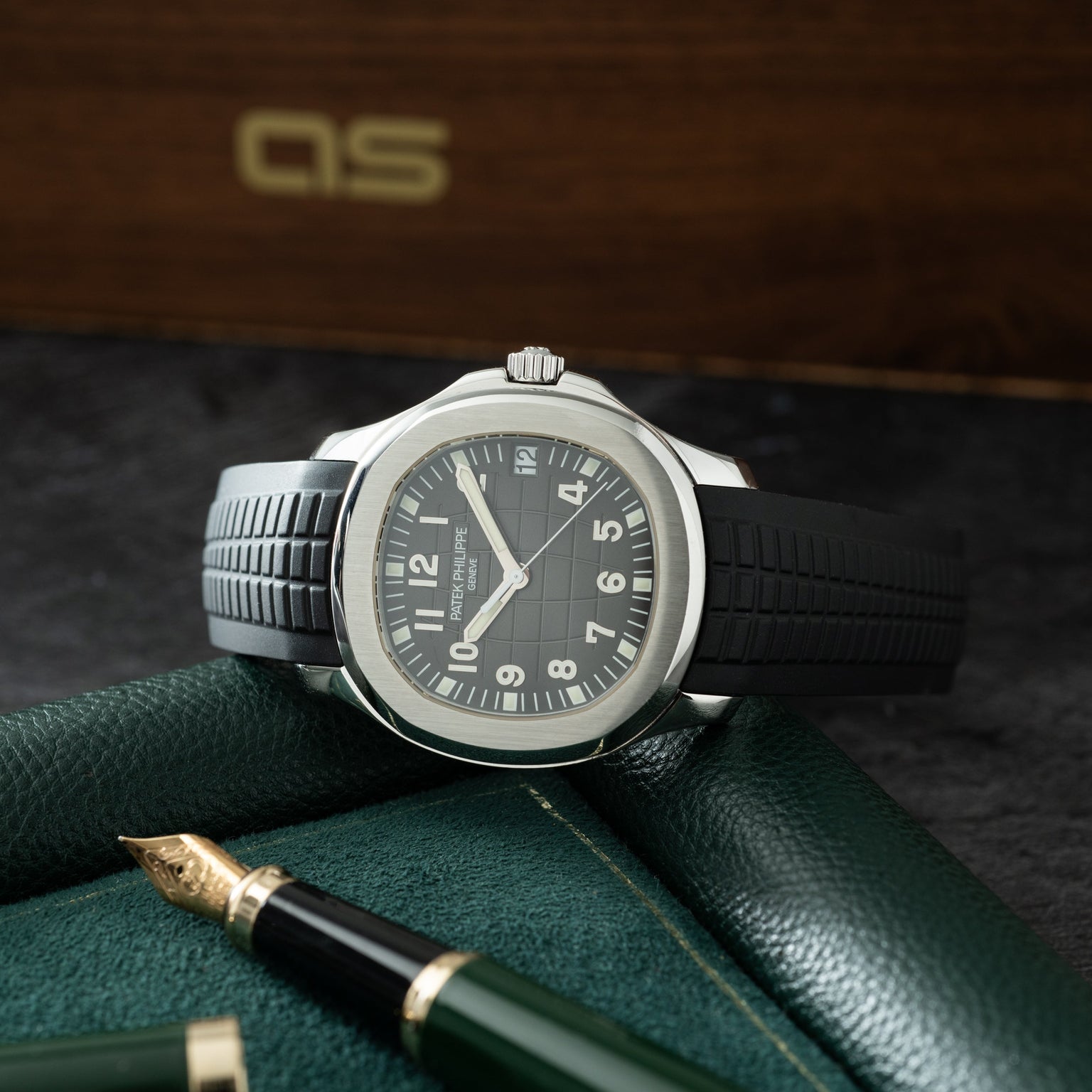 Stainless steel watch with dark dial, white hands and numerals, black rubber strap, displayed on green leather with fountain