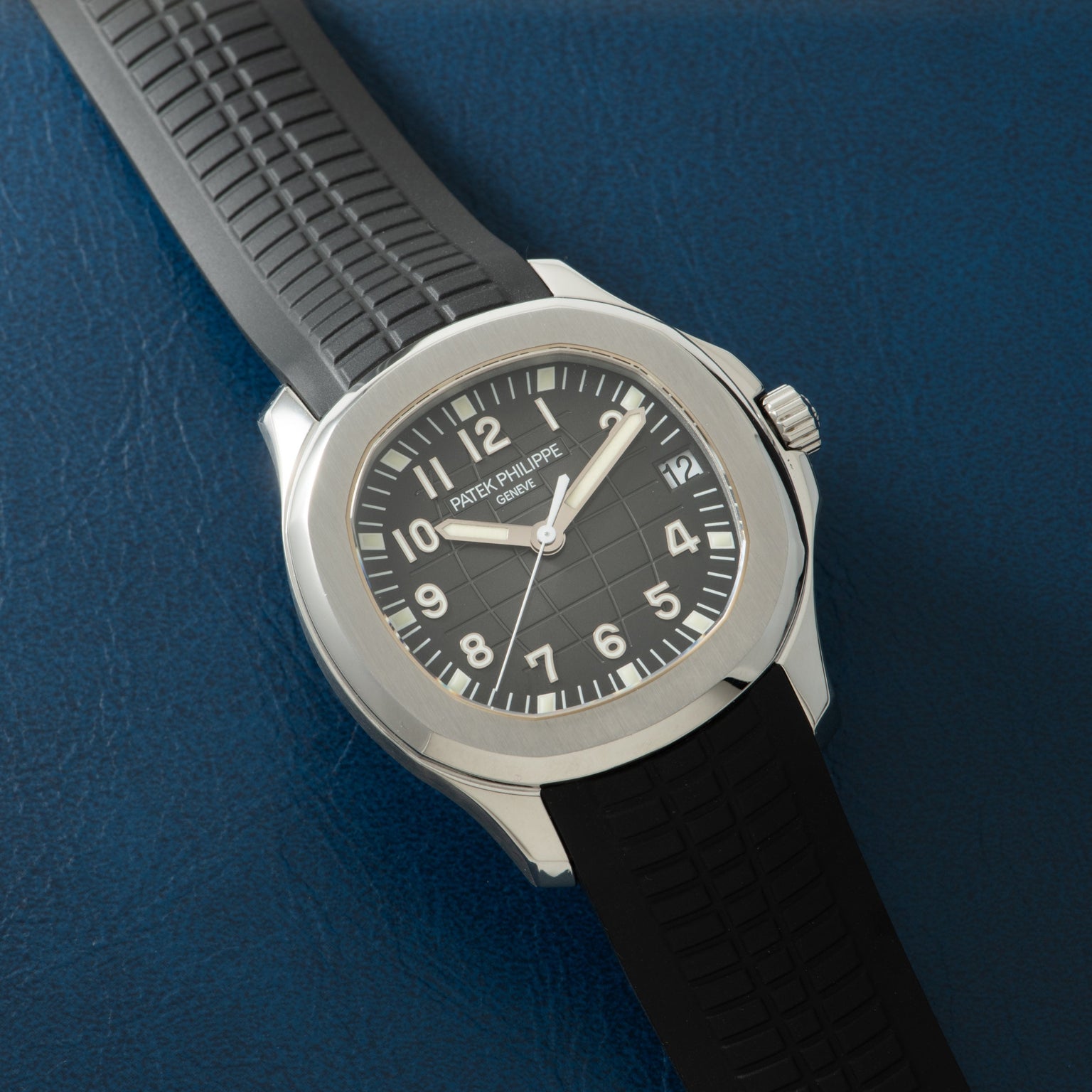 Stainless steel watch with black dial, Arabic numerals, and black rubber strap on blue textured background.