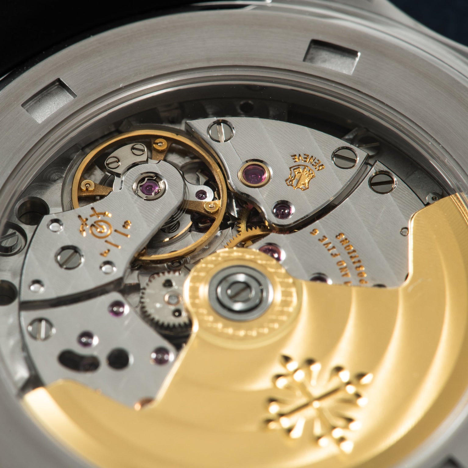 Close-up of watch movement showing gold gears, jewels, and engraved metal plates with Patek Philippe logo.