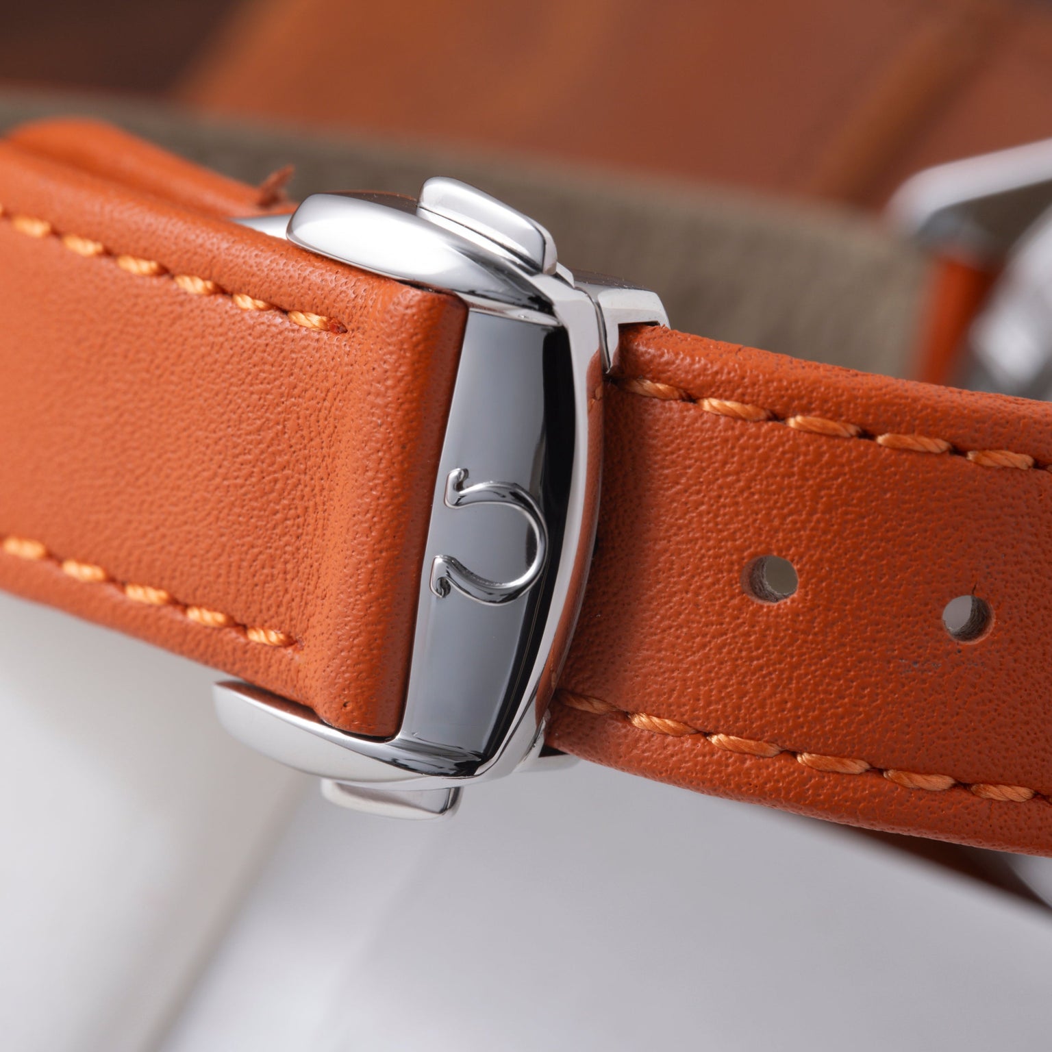 Polished steel watch case with crown detail on orange leather strap with tan stitching.