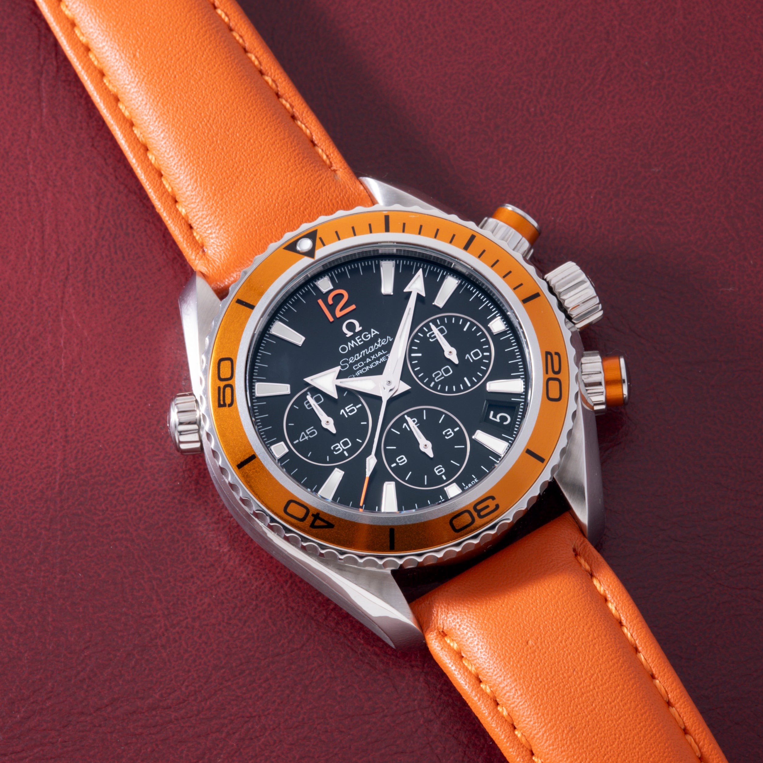 Stainless steel chronograph watch with black dial, orange bezel, and matching orange leather strap on burgundy background.