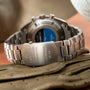 Titanium chronograph watch with blue dial and metal bracelet, shown from rear view on wood surface.