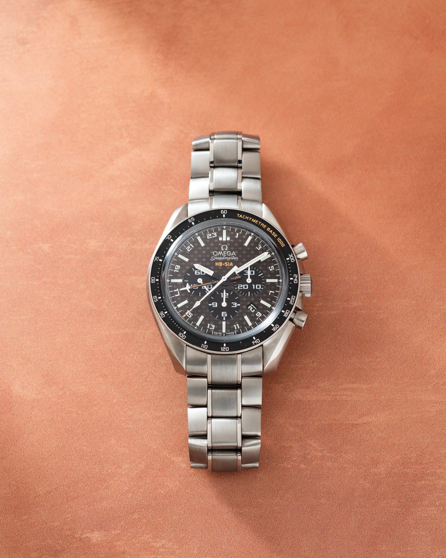 Silver-tone Omega Speedmaster chronograph watch with black dial, tachymeter bezel, and metal bracelet on tan background.
