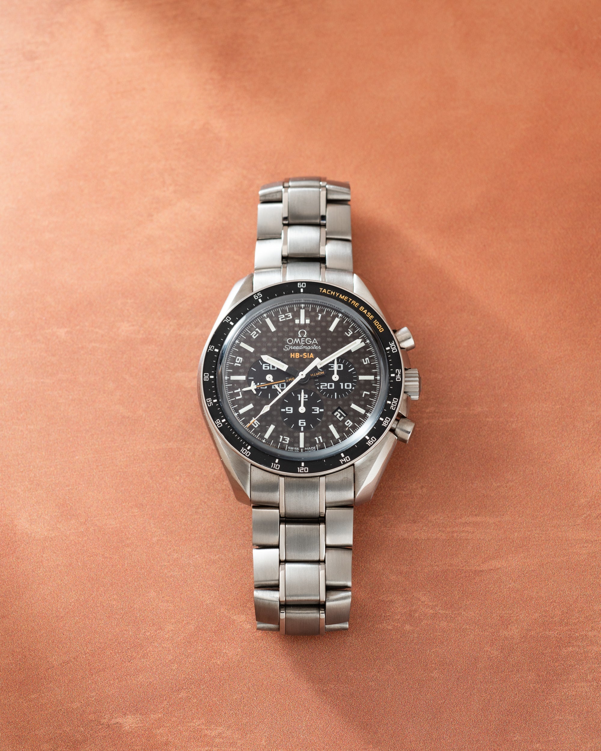 Silver-tone Omega Speedmaster chronograph watch with black dial, tachymeter bezel, and metal bracelet on tan background.