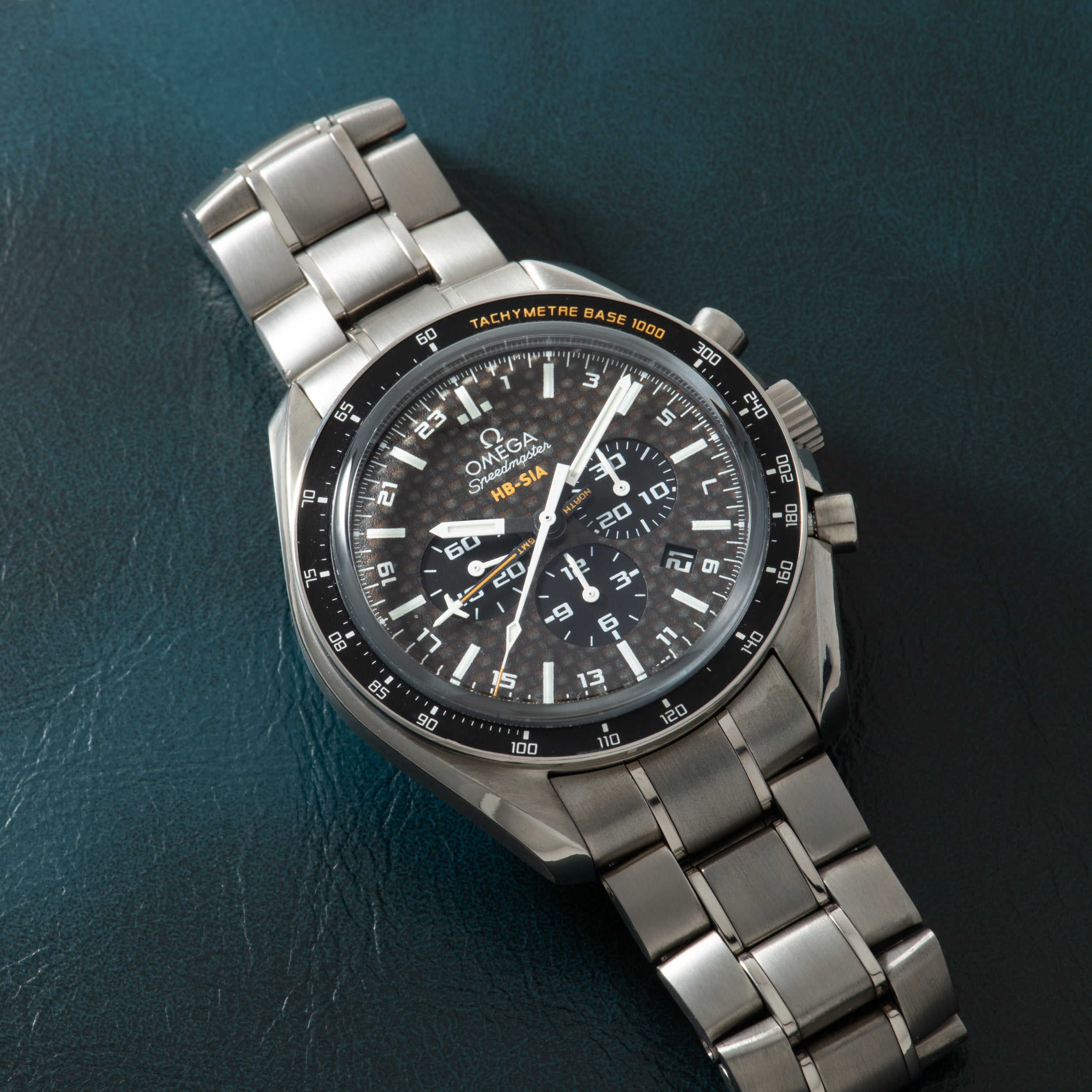 Omega Speedmaster chronograph watch with titanium bracelet, black dial, tachymetre bezel, and three subdials on teal backgrou