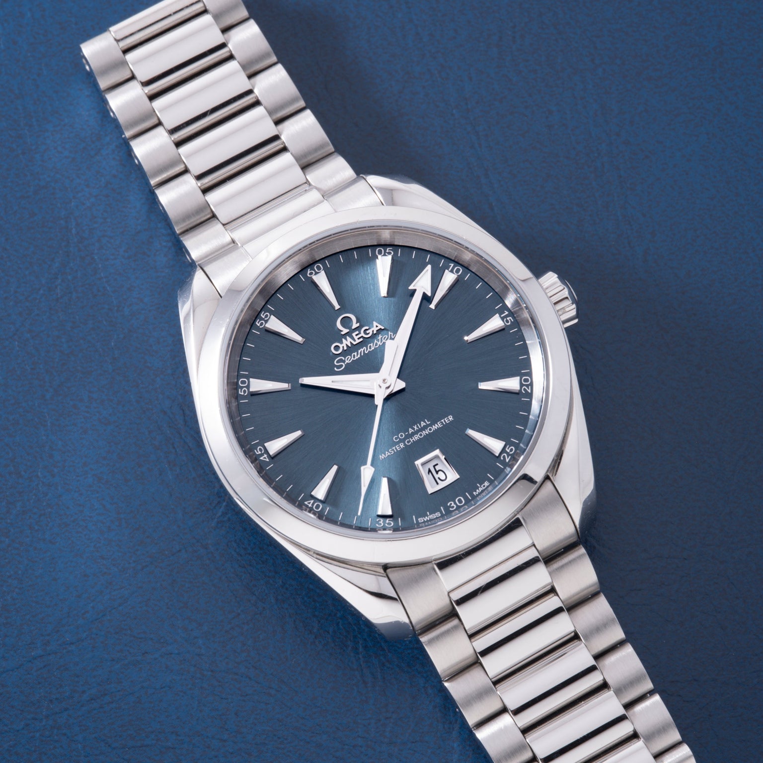 Stainless steel watch with blue dial, white hour markers, date window, and matching metal bracelet on blue background.